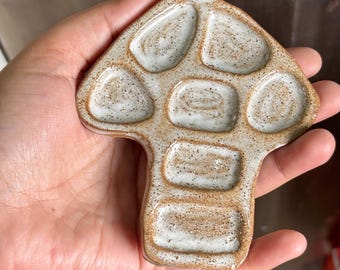 Puede incluir: Una paleta de pintura de cerámica con forma de hongo que presenta un esmalte blanquecino moteado con un borde de arcilla marrón texturizado. La paleta incluye siete pocillos empotrados distintos para mezclar pintura, dispuestos en un diseño simétrico.