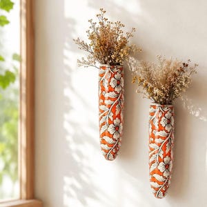 Pode incluir: Dois vasos de parede cilíndricos de cerâmica com um fundo laranja vibrante e um padrão branco de flores e vinhas. Cada vaso está preenchido com um ramo de flores silvestres secas bege, criando uma decoração montada numa parede lisa.