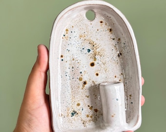 May include: An arched ceramic wall sconce with a speckled off-white glaze featuring brown, gold, and dark blue flecks. The piece has a flat back, a small circular hole at the top for hanging, and a cylindrical candle holder attached at the base.