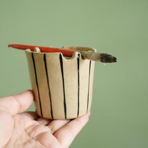 May include: A beige ceramic brush holder with vertical black stripes. A red paintbrush and a paint brush are resting in the holder. The holder is held in a hand against a green background.