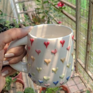 May include: White ceramic mug decorated with colorful heart-shaped embellishments. The mug has a curved handle and is held in a person's hand. The mug is surrounded by plants and a metal fence.