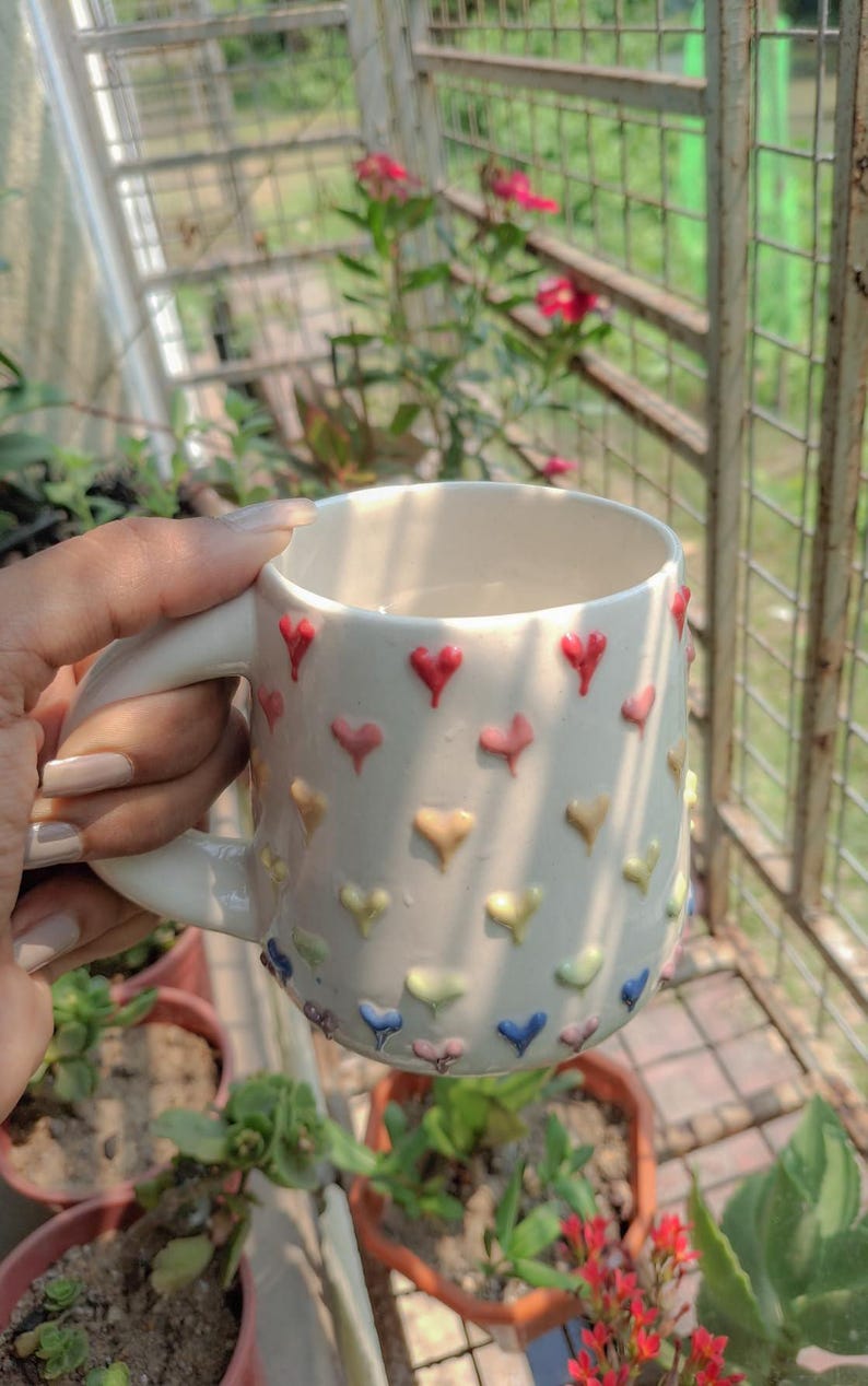 May include: White ceramic mug decorated with colorful heart-shaped embellishments. The mug has a curved handle and is held in a person's hand. The mug is surrounded by plants and a metal fence.