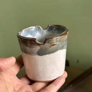 Może przedstawiać: Ręcznie robiona ceramiczna popielniczka. Popielniczka ma białą podstawę, szaro-niebieski pas i brązową krawędź. Wnętrze jest jasnoniebieskie. Ma zakrzywioną krawędź do odkładania papierosów.
