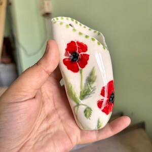 Może przedstawiać: Biały ceramiczny wazon ścienny z ręcznie malowanymi czerwonymi makami z czarnymi środkami i zielonymi łodygami. Krawędź ozdobiona jest serią małych zielonych kropek, a przedmiot ma wydłużony, zakrzywiony kształt z otworem u g&oacute;ry.