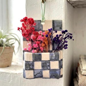 May include: A ceramic wall planter with a cream and navy blue chequered design, holding vibrant dried flowers. The rectangular planter has a curved base and is hung by a green leather strap from a brass hook. The flowers are in shades of red, pink, orange and purple.