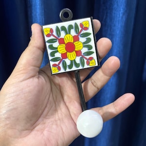 Peut inclure: Un carreau décoratif carré avec un motif floral rouge, jaune et vert, fixé à une poignée noire et une boule blanche. Le carreau a un cadre en métal noir et une petite boucle en haut pour accrocher.