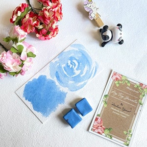Puede incluir: Dos pastillas de pintura acuarela azul sobre una superficie blanca con una pintura de acuarela de una rosa azul. La pintura está en una tarjeta blanca con el texto "My Toy Journalce" y "Handmade Watercolor Journals & Paints" escrito en ella.