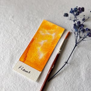 May include: A watercolor painting sample labeled "Flame" with a vibrant orange and yellow gradient. A paintbrush and a sprig of blue flowers are next to the sample on a white textured surface. The painting is a rectangular shape.