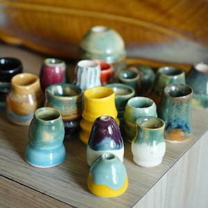May include: A collection of small, handmade ceramic vessels in various shapes and sizes. The pieces feature a diverse range of glazed finishes, including solid yellow, deep purple, teal, blue, white, and multi-coloured blends with glossy textures.