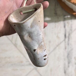 May include: A cone-shaped ceramic wall planter with a speckled beige and white glaze. The vessel features a wide, open top with two small circular holes near the rim, through which a thin, light-colored cord is threaded for hanging purposes.