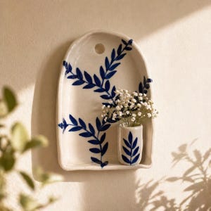 Può includere: Una tasca da parete in ceramica bianca con un design a foglie blu. La tasca ha un piccolo vaso attaccato, contenente fiori bianchi. Il design è un motivo botanico stilizzato. La tasca da parete è montata su una parete di colore neutro.