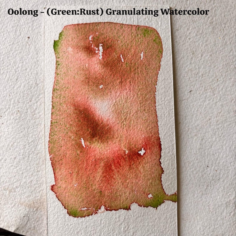 May include: A watercolor swatch on white paper, showcasing a blend of rust, green, and red hues. The text "Oolong - (Green:Rust) Granulating Watercolor" is at the top. The paint has a textured, granular appearance.