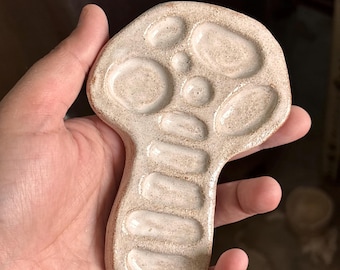 May include: A mushroom-shaped ceramic palette with a speckled beige finish. The surface features multiple recessed circular and oval indentations for holding paint, with a smooth, glossy glaze covering the entire top side of the object.