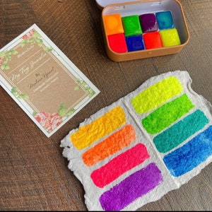 May include: A set of eight watercolor paints in a metal tin. The paints are arranged in two rows of four, with each paint a different color. The colors are yellow, green, purple, blue, orange, red, pink, and a bright neon yellow. The tin is labeled "My Tiny Journal" and "Handmade Watercolor Journal & Paints".