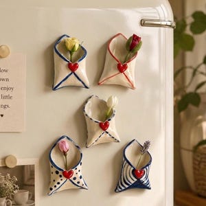 Può includere: Cinque vasi da parete in ceramica a forma di busta attaccati a un frigorifero. Ogni vaso bianco presenta un piccolo cuore rosso e contiene un singolo fiore o rametto. Le buste hanno vari motivi dipinti in blu o rosso, tra cui righe e pois.