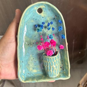 Może przedstawiać: Ścienny wazon ceramiczny z łukowatą płytką tylną w nakrapianym turkusowym szkliwie. Przymocowana kieszonka ma strukturę plastra miodu i zawiera bukiet suszonych kwiatów w żywych odcieniach jaskrawego różu i głębokiego błękitu.