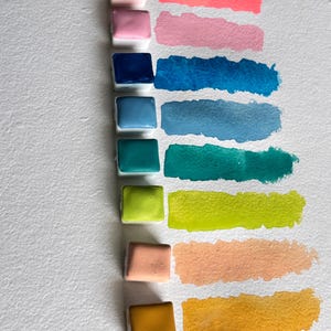 May include: A collection of watercolor paint pans in various colors, including pink, blue, green, and yellow, with corresponding swatches. The pans are arranged vertically on white paper, showcasing the range of hues and their application.