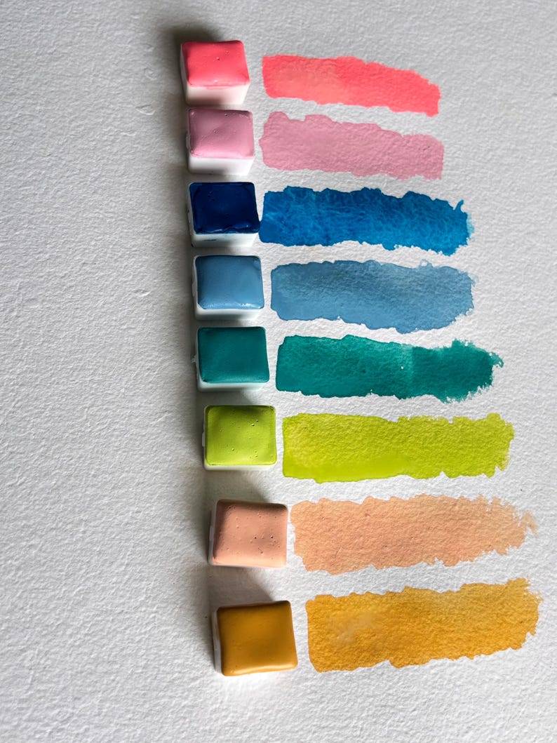 May include: A collection of watercolor paint pans in various colors, including pink, blue, green, and yellow, with corresponding swatches. The pans are arranged vertically on white paper, showcasing the range of hues and their application.