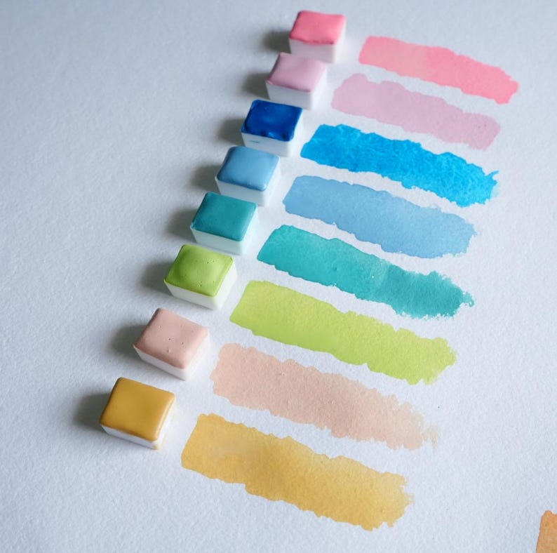May include: A collection of watercolor paint cubes in various colors, including pink, blue, green, and yellow, with corresponding paint swatches. The cubes are arranged diagonally on a white surface, showcasing the range of hues and their application.