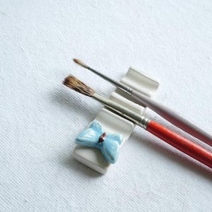 Puede incluir: Dos pinceles descansan sobre un soporte de cer&aacute;mica blanco con un lazo azul claro. Un pincel tiene un mango rojo, el otro un mango morado. Los pinceles est&aacute;n sobre un fondo de lienzo blanco.