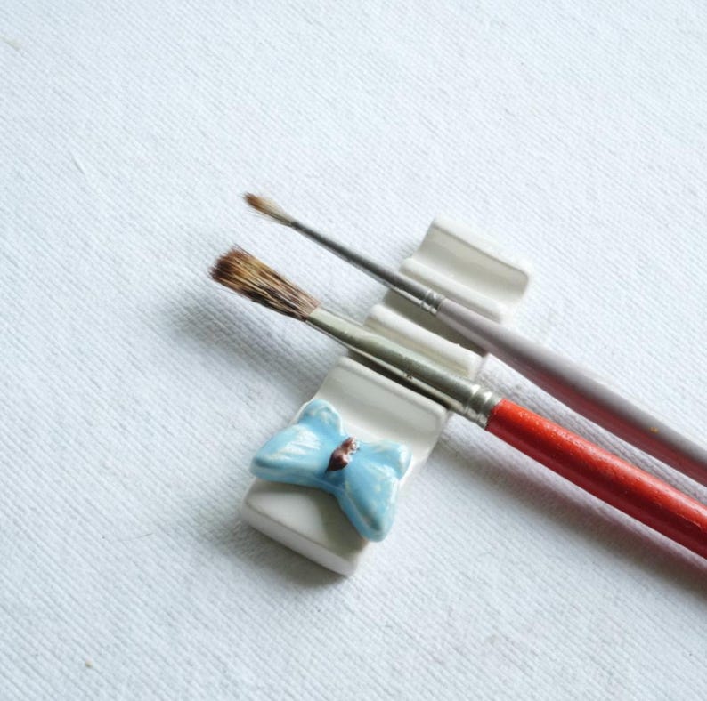 Puede incluir: Dos pinceles descansan sobre un soporte de cer&aacute;mica blanco con un lazo azul claro. Un pincel tiene un mango rojo, el otro un mango morado. Los pinceles est&aacute;n sobre un fondo de lienzo blanco.