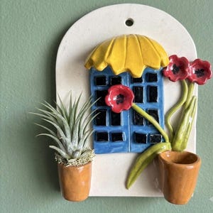 May include: A ceramic wall plaque featuring a white arched base with a blue building facade, a yellow awning, and black windows. Two small brown pots are attached, one holding a green air plant and the other containing three red flowers.