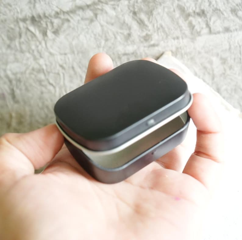 May include: A small, rectangular black tin with a hinged lid, held open in a hand. The interior of the tin is a light silver color. The tin is empty and appears to be new.