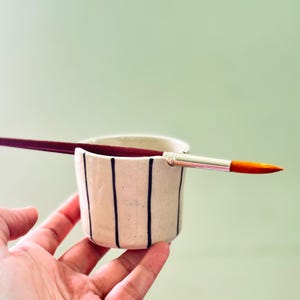Puede incluir: Un pequeño vaso de cerámica color blanco hueso con rayas negras verticales y un borde con muesca diseñado para sostener un pincel. Un pincel largo con mango rojo oscuro, virola plateada y punta naranja descansa horizontalmente en la muesca.