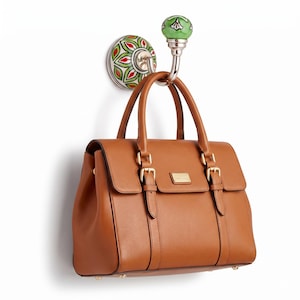 Könnte beinhalten: Eine braune Lederhandtasche mit goldenen Schnallen hängt an einem dekorativen Wandhaken. Der Haken hat einen grünen und silbernen Knauf und eine Keramikbasis mit einem rot-grün-weißen Blumenmuster.