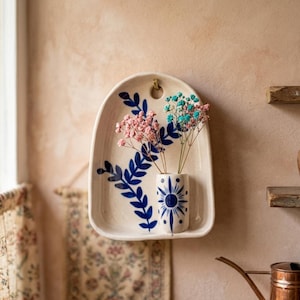 Może przedstawiać: Kremowa ceramiczna kieszeń ścienna z łukowatym szczytem i otworem do zawieszenia. Przedmiot posiada ręcznie malowany wzór ciemnoniebieskich liści oraz mały, przymocowany cylindryczny wazon ozdobiony niebieskim motywem promieni słonecznych z kwiatami.