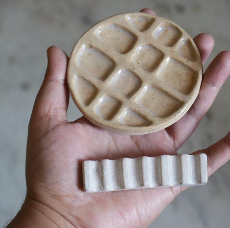 May include: A round, tan ceramic paint palette with multiple recessed wells and a separate rectangular, off-white ceramic brush rest featuring a series of vertical ridges, both with a smooth, glossy finish and visible speckled texture.