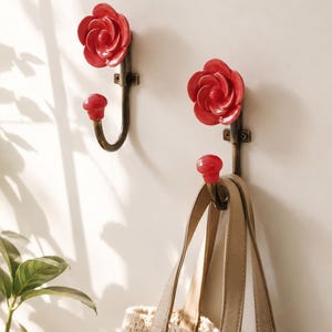 May include: Two red rose-shaped wall hooks with a bronze finish. One hook holds a beige tote bag. The hooks are mounted on a white wall, with a plant in the lower left corner. The hooks are decorative and functional.