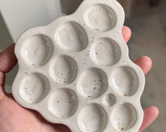 Puede incluir: Una paleta de pintura de cerámica color blanco roto con una forma orgánica irregular que presenta diez pocillos circulares de distintos tamaños. La superficie tiene un acabado brillante con pequeñas motas marrones dispersas.