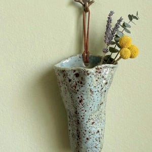 May include: A wall-mounted ceramic vase with a light blue glaze and brown speckles, suspended by a brown leather cord from a small metal hook. The vase contains a small arrangement of dried lavender sprigs and yellow billy ball flowers.