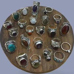 Peut inclure: Une collection de bagues en argent avec diverses pierres précieuses, dont l'améthyste, la turquoise et l'onyx, exposées sur une surface en bois. Les bagues présentent des designs variés, des bandes simples aux montures ornées, affichant des formes et des couleurs diverses.