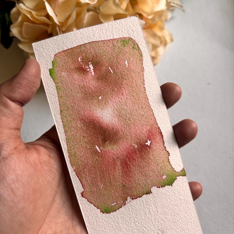 May include: A rectangular watercolor swatch on textured paper. The artwork features a blend of pink and brown hues with green accents along the edges. The paper is held by a hand, with a blurred floral arrangement in the background.