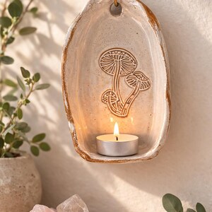 Pode incluir: Um porta-velas de cerâmica montado na parede com um design de cogumelos. O suporte branco sujo tem uma borda marrom e uma vela de chá acesa dentro. Um gancho de latão é fixado para pendurar. Cristais e vegetação em primeiro plano.