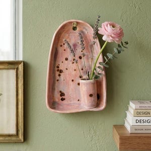 Pode incluir: Um recipiente de cerâmica para parede com formato arqueado e esmalte manchado em tons de rosa e castanho. Inclui um pequeno suporte cilíndrico integrado contendo ramos de alfazema e uma flor de ranúnculo cor-de-rosa.