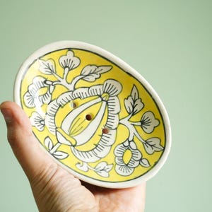 Op de afbeelding: Een gele ovale keramische zeepbak met een wit bloemenmotief. De zeepbak heeft afvoergaatjes.