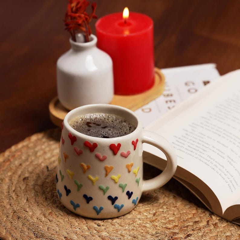 May include: A white ceramic mug filled with coffee, adorned with colorful heart-shaped embellishments. A red candle burns beside a small white vase with dried flowers. An open book rests on a woven mat, creating a cozy, inviting scene.