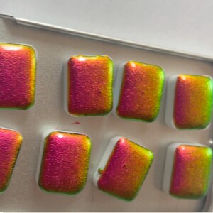 K&ouml;nnte beinhalten: Eine Palette mit acht rechteckigen Aquarellfarben. Die Farben haben einen metallischen Glanz, wobei die Farben von Pink zu Gold und Gr&uuml;n wechseln. Die Farbt&ouml;pfe befinden sich in einer kleinen, silberfarbenen Metallbox.