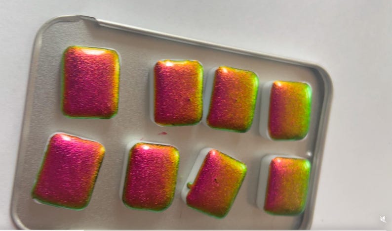 K&ouml;nnte beinhalten: Eine Palette mit acht rechteckigen Aquarellfarben. Die Farben haben einen metallischen Glanz, wobei die Farben von Pink zu Gold und Gr&uuml;n wechseln. Die Farbt&ouml;pfe befinden sich in einer kleinen, silberfarbenen Metallbox.
