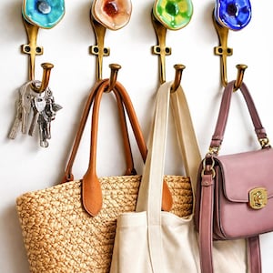 May include: Four decorative wall hooks with colourful floral designs in blue, orange, green, and blue. The hooks are holding a straw tote bag, a canvas tote bag, a pink purse, and a set of keys. The hooks are made of brass.