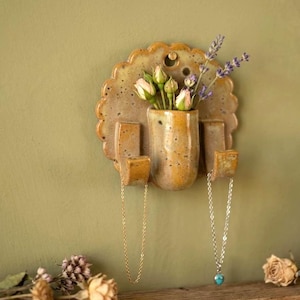 May include: A wall-mounted ceramic organiser with a scalloped edge, speckled mustard-yellow glaze, and a central cylindrical vase holding small roses and lavender. Two curved hooks extend from the base, each supporting a delicate metal chain necklace.