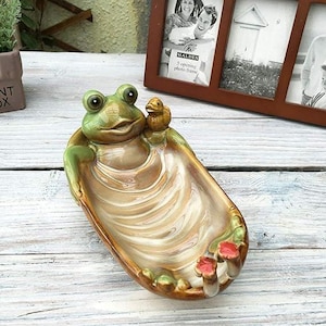 Może przedstawiać: Ceramiczna miska w kształcie żaby z figurką kaczki. Miska jest głównie beżowa z zielonymi i brązowymi akcentami. Żaba ma duże, okrągłe oczy i trzyma miskę. Miska ma wzór muszli.