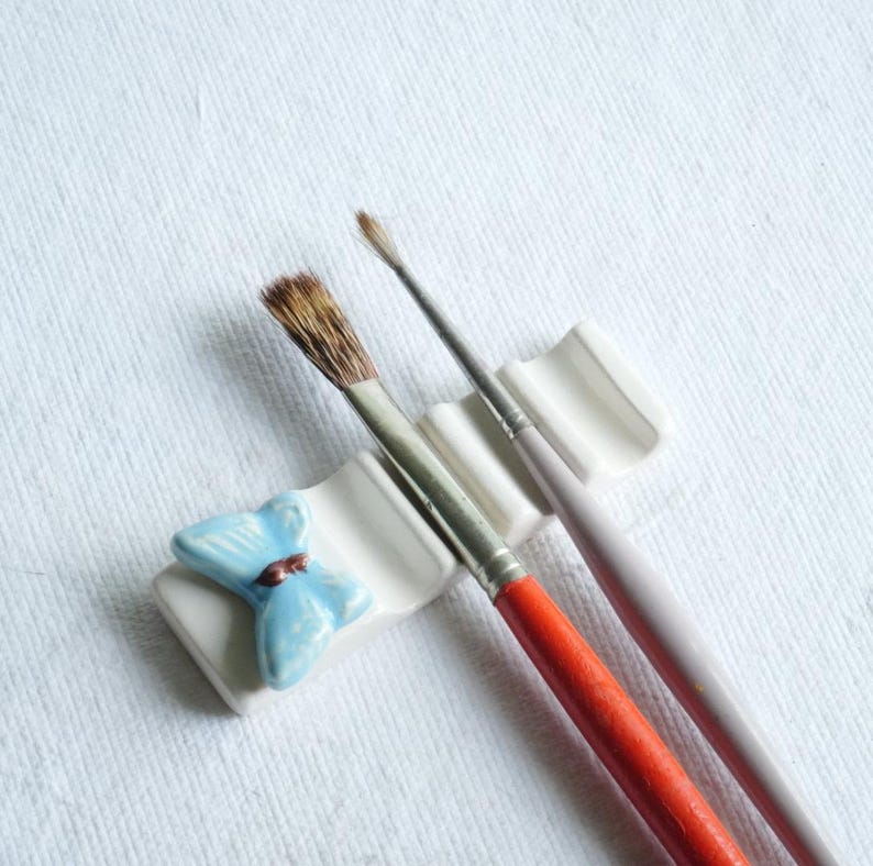Puede incluir: Un soporte de cer&aacute;mica blanco para pinceles con un dise&ntilde;o de mariposa azul, que sostiene dos pinceles. Un pincel tiene un mango rojo, el otro un mango rosa p&aacute;lido. Los pinceles tienen cerdas marrones.