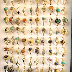 Op de afbeelding: Een verzameling goudkleurige metalen ringen gerangschikt in een raster, met diverse edelstenen zoals turkoois, tijgeroog, maansteen en lapis lazuli, naast ingewikkelde filigraan, halve maan en geometrische metalen ontwerpen.