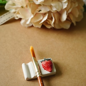 May include: A white ceramic paintbrush holder with a red strawberry design, holding a paintbrush with a wooden handle and yellow bristles. A bouquet of cream-colored flowers is in the background.