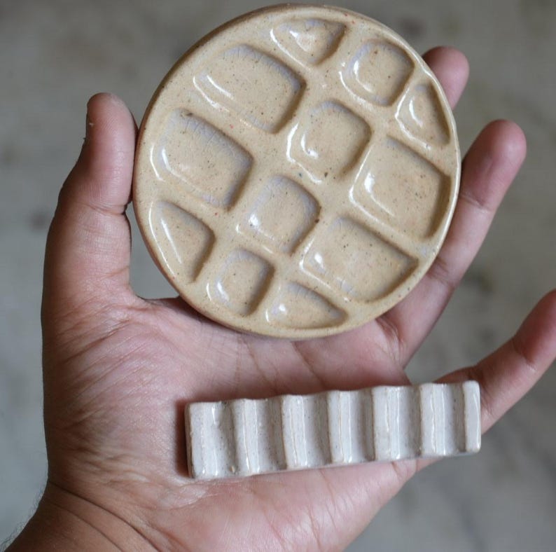 May include: A round, beige ceramic palette with a grid of recessed square wells and a separate rectangular, off-white ceramic brush rest featuring a series of vertical ridges. Both items have a smooth, glossy finish and speckled texture.