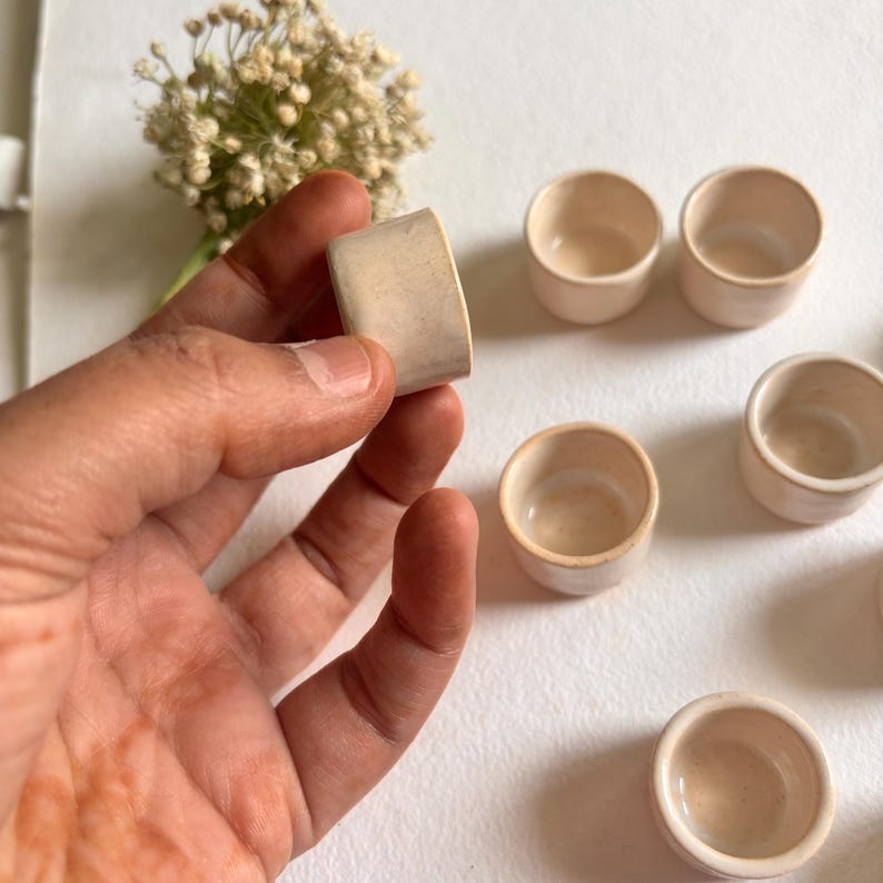 May include: Set of six small, white ceramic cups with a slightly textured surface. The cups are arranged in a circle on a white background.